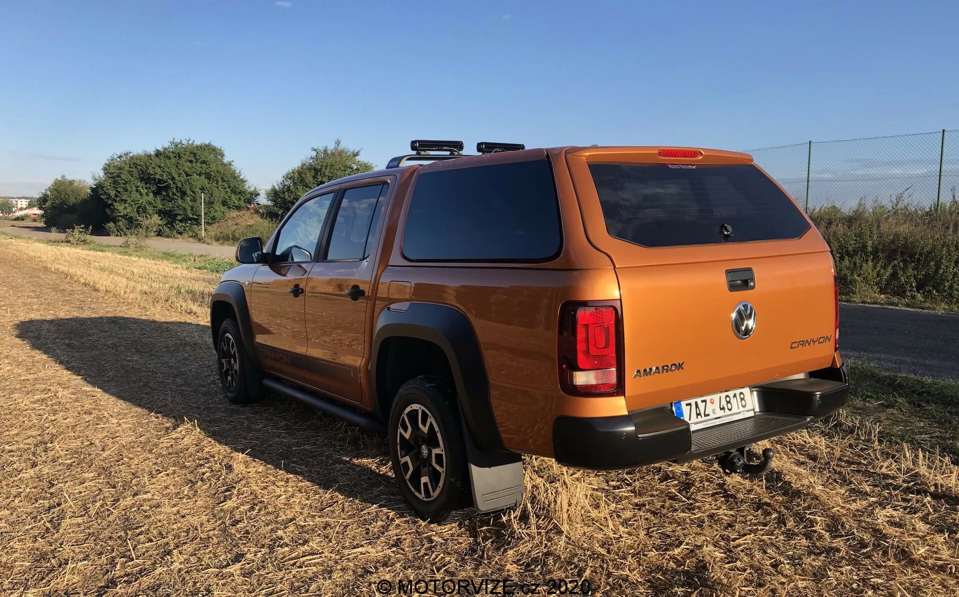 Bronz vagy égett narancs színű modern haszongépjármű vagy terepjáró jármű hátsó negyed nézete, amely feltehetően közepes méretű SUV vagy pick-up, figyelembe véve a rakodótér fedelét. A szög kiemeli a hátsó részt és az oldalsó profilt, beleértve a hátsó kereket és az utasajtót, terepen, háttérben növényzettel. A jármű robusztus, szögletes kialakítással rendelkezik, ami tipikus a terepjárókhoz, körbeérő hátsó lámpákkal, nagy, terepre alkalmas gumiabroncsokkal fekete alufelniken, látható emblémákkal, tetősínekkel, hátsó sötétített üvegekkel és szilárd rakodótér fedéllel.