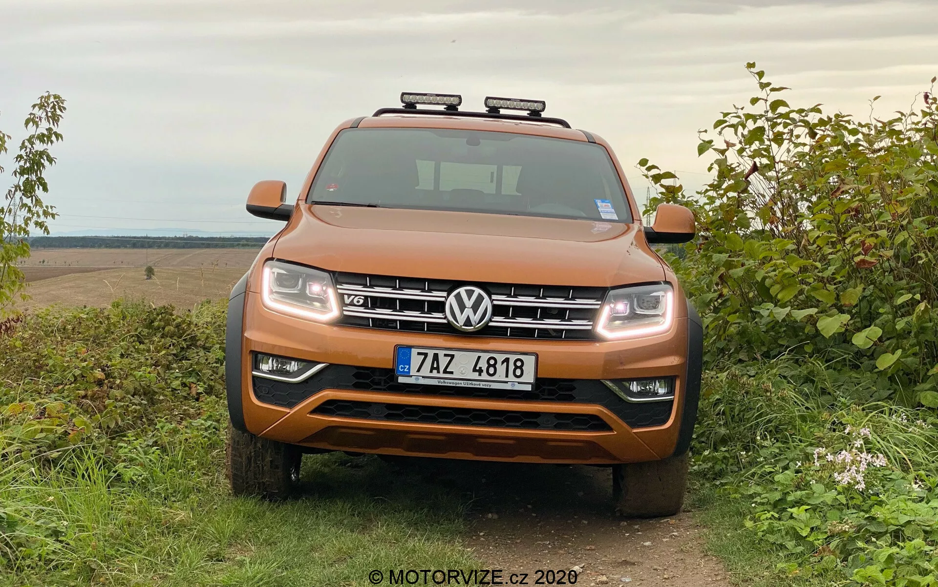 Első nézet egy narancsmetál Volkswagen Amarok SUV-ről, amely füvön parkol vidéki off-road háttérrel, szimmetrikus hűtőrácsot, fényszórókat, króm díszítéseket és kiegészítő tetőlámpákat mutat.
