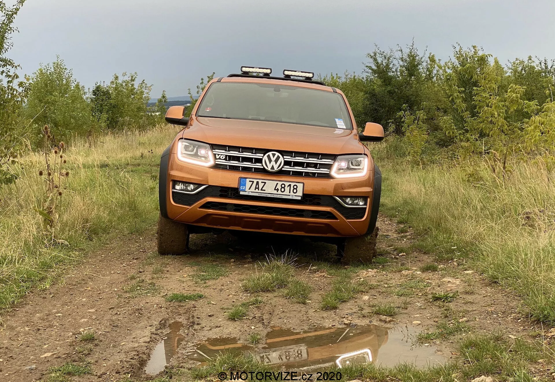 Elölnézeti kép egy fémes narancssárga Volkswagen Amarok 2016 SUV-ról egy egyenletlen terepen, háttérben fűvel és cserjékkel. A jármű enyhe lefelé irányuló szögben néz a kamerára, az előtérben egy sekély pocsolyával, amely visszatükrözi az autó képét. Az első rács közepén a Volkswagen embléma, a motorháztető pedig bemutatja az ő vonalait és formáit. A modern LED fényszórókkal és az alatta elhelyezkedő ködlámpákkal felszerelt, az alsó részen található ezüst csúszólemez terepjáró-képességekre utal. Kiegészítő lámpák vannak a tetőcsomagtartóra szerelve, növelve a jármű kalandos, robusztus megjelenését. A rendszámtábla az első lökhárító közepén található. A kép teljes szélességében ábrázolja a járművet, bemutatva a technológiával, formatervezéssel és terepjáró-alkalmazkodással kapcsolatos részleteket.