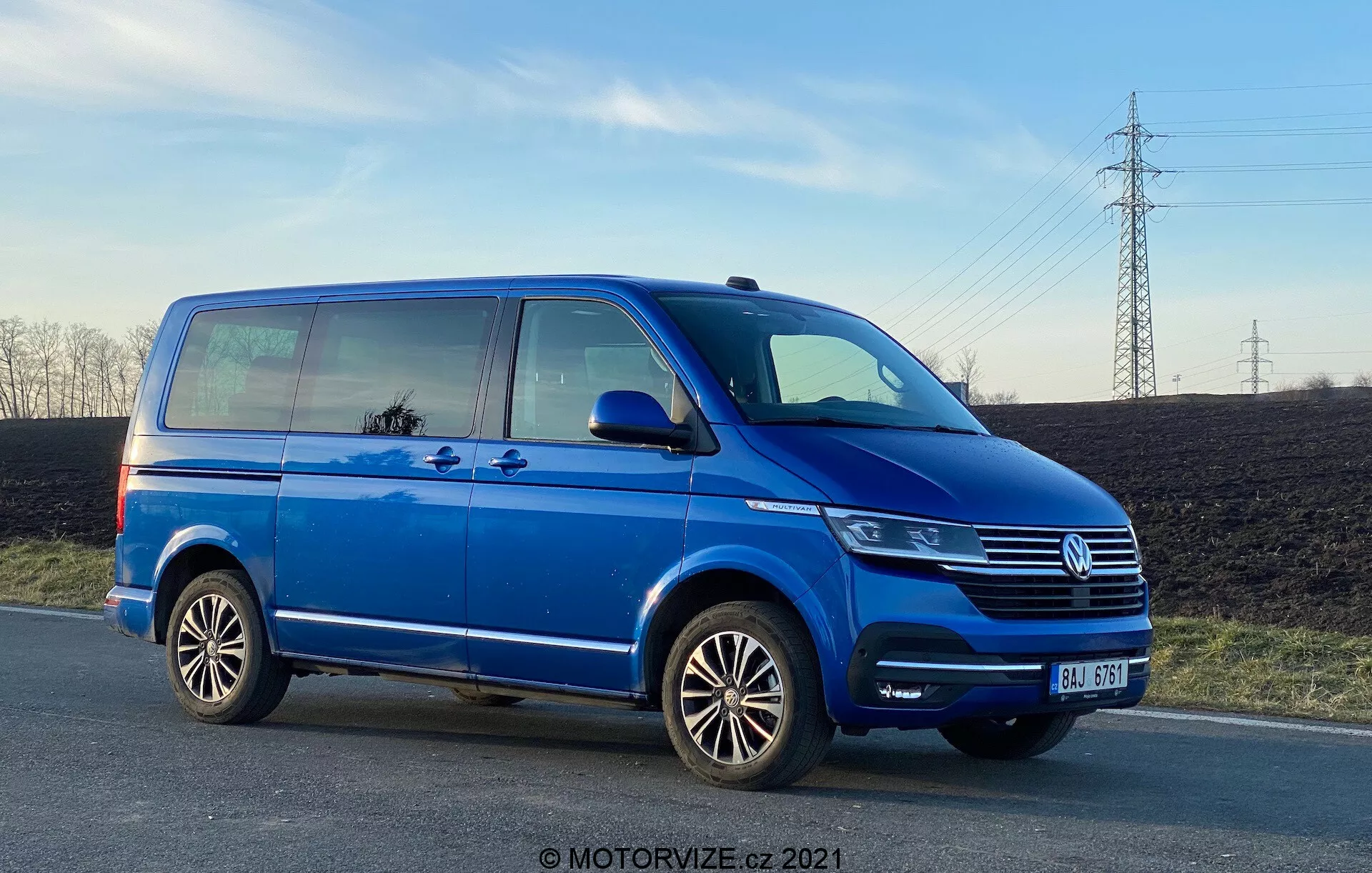 Egy kék Volkswagen Transporter T6 furgon, amely alacsony szögből mutatja oldalsó profilját, kihangsúlyozva annak hosszát és magasságát. A kép részletei közé tartozik a toló utasajtó, az elülső utasajtó, a hátsó utas oldali kerék, a többrudas alumínium felnik, valamint a fényszóró, a Volkswagen rács emblémával és az első lökhárító részleges nézetei. A késő délután készült fotó megmutatja a hasmagasságot, és tükrözi az eget és a környezetet az ablakokban, tiszta ég és elektromos vezetékek háttérrel.
