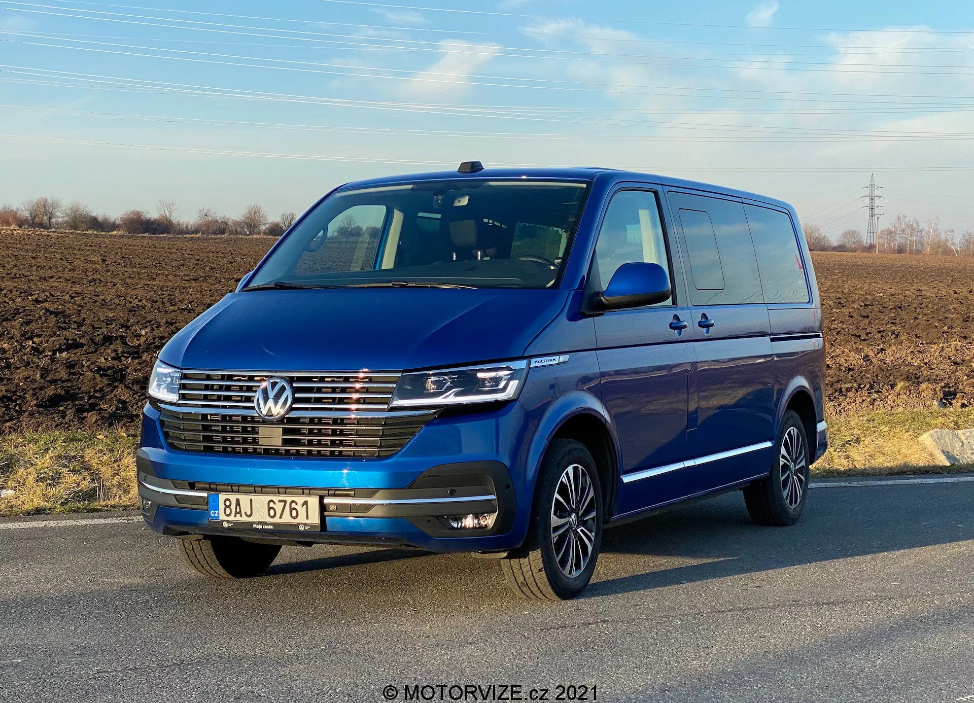 Egy 2019-es kék Volkswagen Transporter T6.1 Multivan negyedik szembenézete tiszta ég alatt. A jármű elején egy Volkswagen embléma látható, LED nappali menetfények, krómozott díszléc az elülső rácson és többrudazott alufelnik. A karosszéria színével azonos tükrök és lökhárítók magas specifikációjú modellt sugallnak. Láthatóak az ajtók és kilincsek, beleértve a hátsó tolóajtót is, ami több utas vagy rakomány szállítására utal.