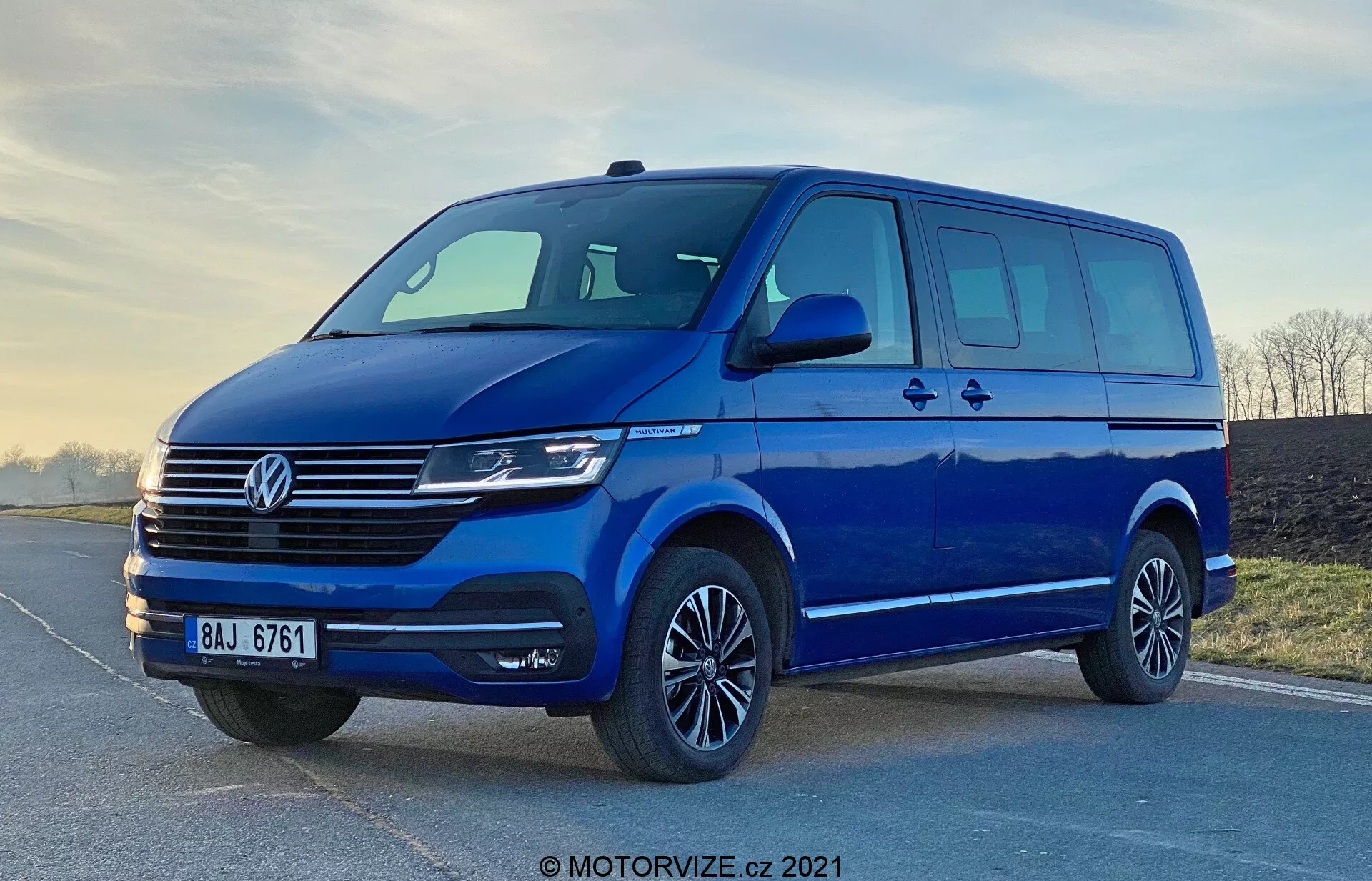 Egy kék 2019-es Volkswagen Transporter T6.1 Multivan kép, alacsony szögből fotózva, hogy kiemelje annak statúráját és méretét. Az elülső háromnegyedes nézet bemutatja a jármű elülső és oldalsó kialakítását, beleértve a jellegzetes Volkswagen hűtőrácsot vízszintes vonalakkal, a központi emblémát, a modern fényszórókat, amelyek valószínűleg LED vagy Xenon technológiát tartalmaznak, és egy elegáns elülső lökhárítót széles légbeömlővel. A beépített irányjelzővel ellátott áramvonalas oldalsó tükrök, a tolóajtóval ellátott nagy ablakfelület, az alacsony profilú gumiabroncsokkal felszerelt könnyűfém felnik, valamint a lágy világítás kiemeli a furgon praktikumát és esztétikai vonzerejét. A 'MOTORVIZE.cz 2021' vízjel azt sugallja, hogy a kép 2021-ben készült.