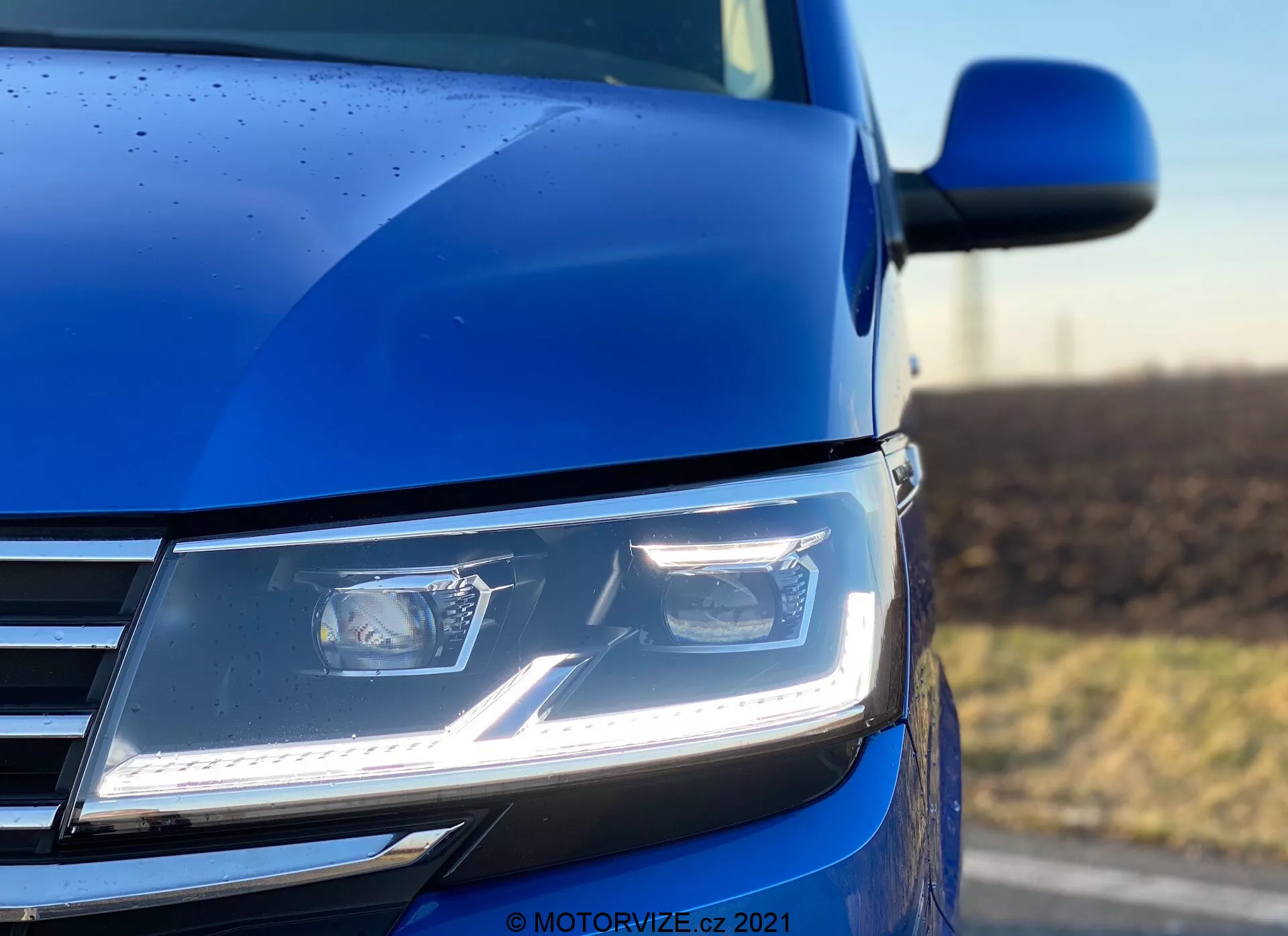 Egy közeli, alacsony szögből készült kép a 2019-es kék VW Nutzfahrzeuge Transporter bal elülső részéről (T6.1 Multivan), amelyen a modern, szögletes LED fényszórók nappali menetfényekkel, a krómozott hűtőrács része, valamint a motorháztető kontúrja és az oldalsó tükör látható. Az alacsony mélységélességgel készített fotó a fényszórót és a környező karosszériát emeli ki, hangsúlyozva az autó fejlett világítástechnikáját, kortárs dizájnját és a festék minőségét, melyet az esőcseppek hangsúlyoznak, jelezve az autó részletes külső bevonatát.