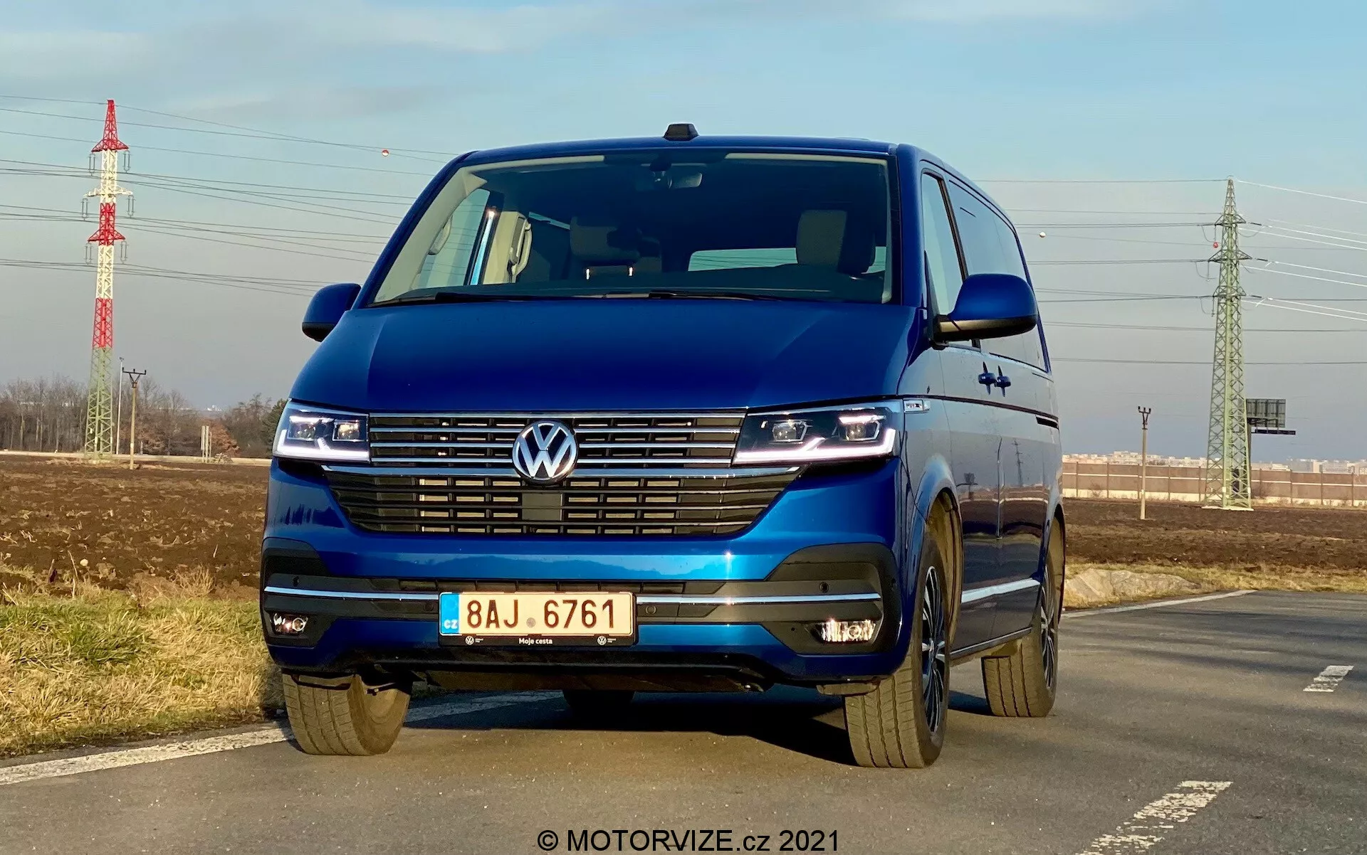 Kék Volkswagen T6-1 Multivan 2019 alacsony elülső szögből fényképezve, kiemelve a hűtőrácsot, LED fényszórókat nappali menetfényekkel, Volkswagen emblémát, széles hűtőrácsot, kéttónusú lökhárítót és könnyűfém kerekeket; egy út szélén parkol, tiszta égbolttal, távoli növényzettel, villanyoszloppal és az infrastruktúrával a háttérben.