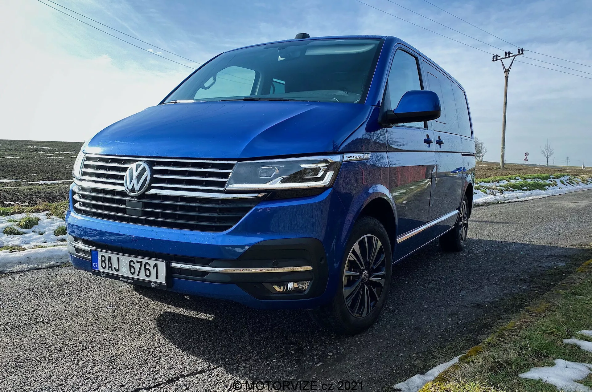 Kék Volkswagen T6.1 Multivan Transporter 2019 alacsony elülső háromnegyedes szögből fényképezve, bemutatva az autó elülső részét, rácsot a prominens Volkswagen logóval, vízszintes krómos rudakkal, szögletes LED fényszórókkal nappali menetfénnyel, első lökhárítót integrált ködlámpákkal és légbeömlőkkel, dinamikus vonalat húzva az autó oldalán, többlengős könnyűfém keréktárcsákat, integrált irányjelzőket az oldalsó tükrökben, sötétített üvegeket. A jármű egy nedves úton parkol havas fű mellett, vidéki háttérrel, amely tartalmazza a közműoszlopokat és a nyitott eget.