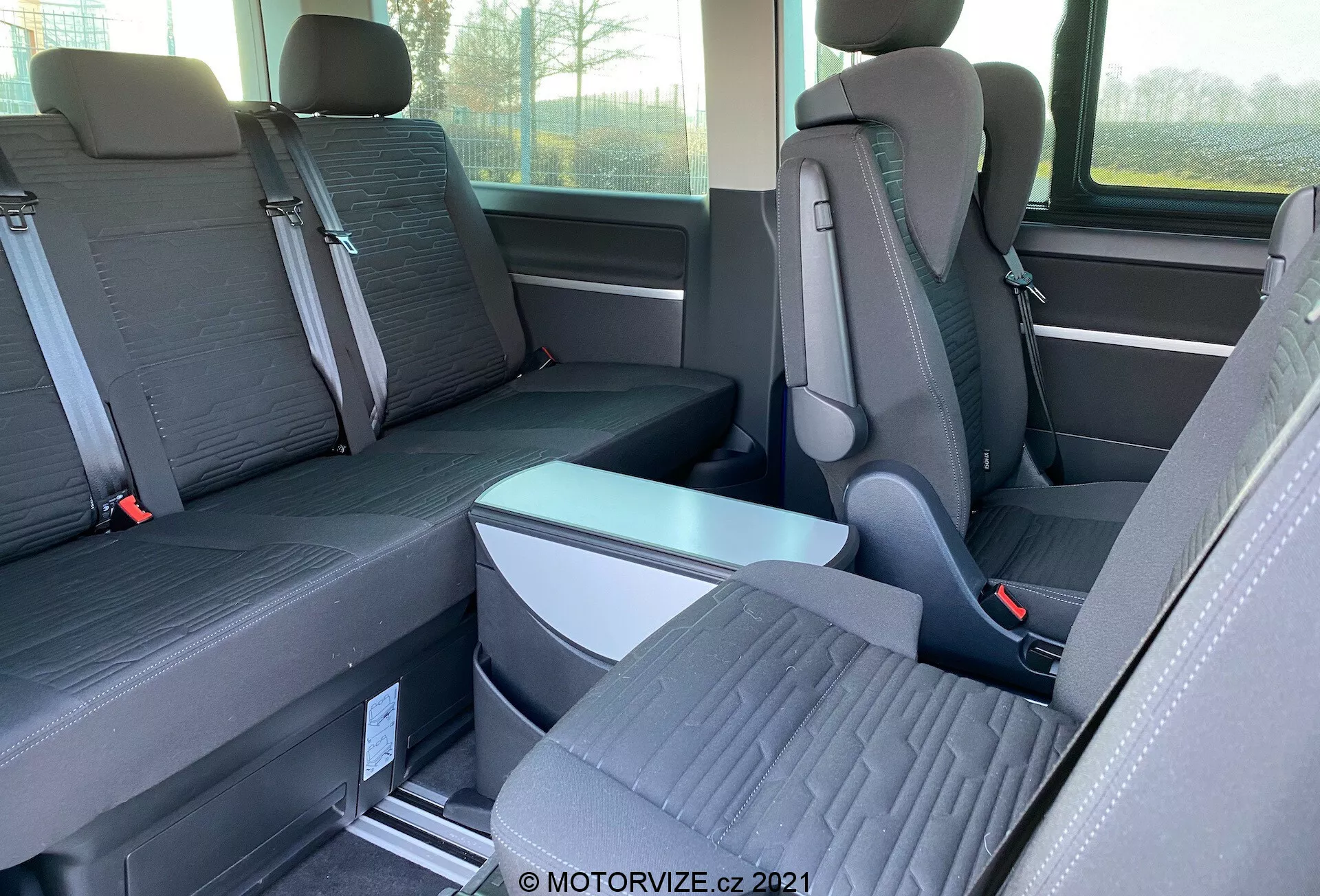 VW-Nutzfahrzeuge Transporter 2019 (T6.1 Multivan) belső nézete, amely a második és harmadik sor üléseire összpontosít. Szemmagasságból, oldalról fényképezve a kép egyéni üléseket mutat kiemelkedő fejtámlákkal a második sorban és egy háromszemélyes padot a harmadik sorban. Az ülések kéttónusú szövetből készültek, és a padlón fém sínek találhatók az átrendezhető ülésekhez. Látható egy lehajtható kartámasz és a biztonsági öv csatjai. Részben látható az ablak és az oldalsó tároló is.