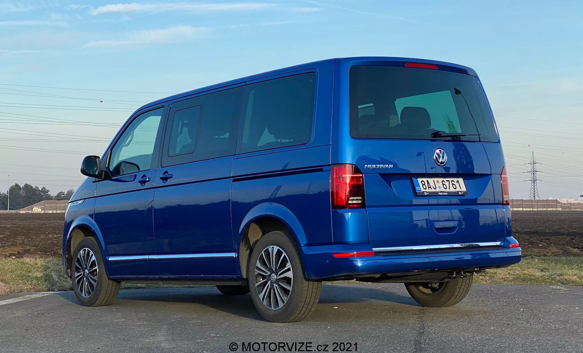 Egy kék VW-Nutzfahrzeuge Transporter 2019 (T6-1 Multivan) látható háromnegyedes hátsó nézetből. A kép megmutatja a hátsó lámpákat, hátsó ablakokat és hátfalat, valamint az oldalsó profilját az utasajtókkal és ablakokkal. A furgon a keret nagy részét kitölti szemmagasságban, feltárva az osztott hátsó ajtókat és több oldalsó ablakot, ami utasmodellt sejtet. Az ötvözet keréktárcsák, a karosszéria színére festett lökhárítók és az oldalsó tükrök magas felszereltségi szintet sugallnak. A megvilágítás nappali, derült égboltú fényviszonyokat jelez, éles árnyékokkal a jármű alatt.