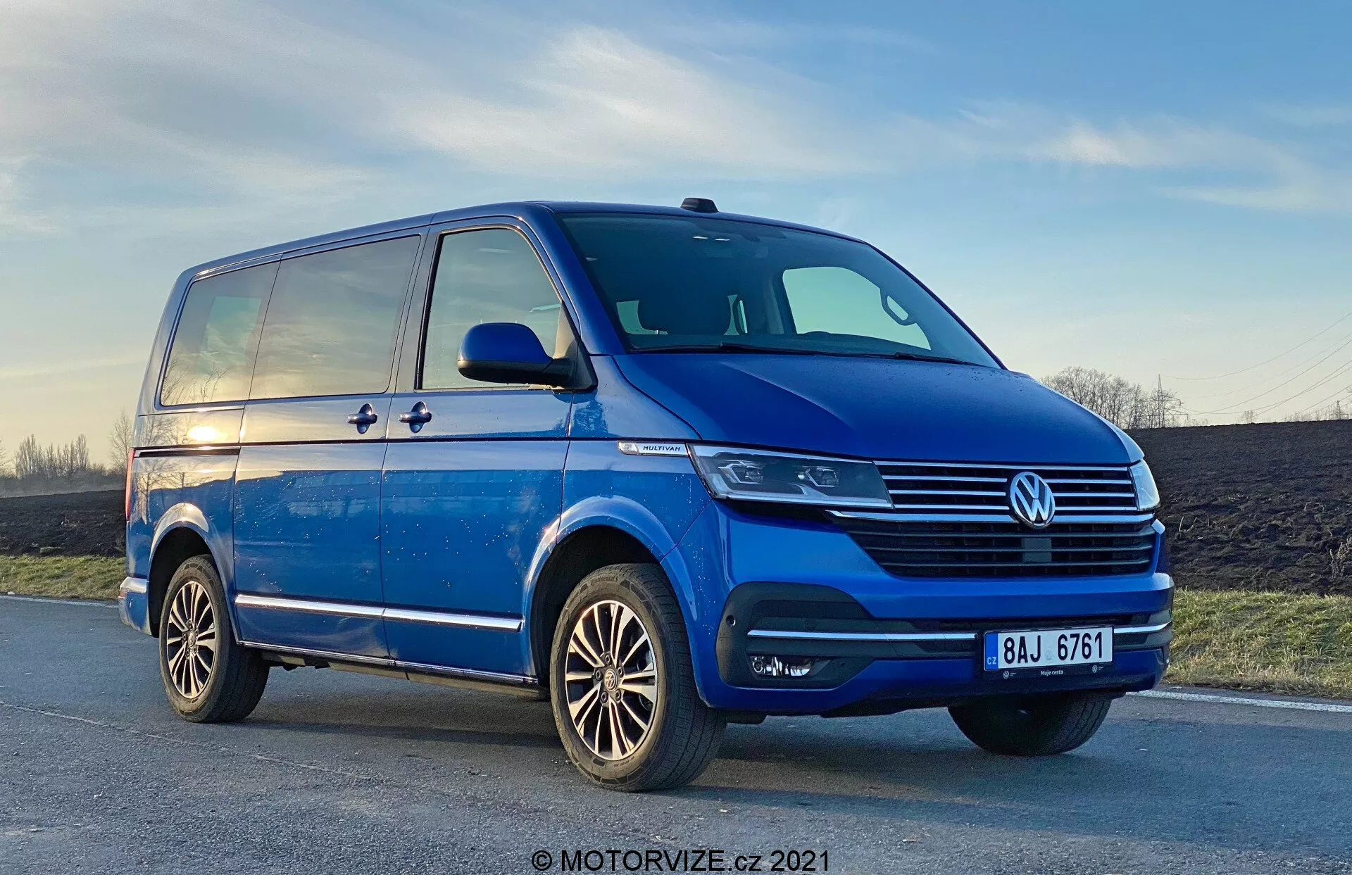 Alacsony szögből készült kép egy 2019-es kék Volkswagen T6-1 Multivanról, amely az első bal kvadráns háromnegyed nézetét mutatja. A furgon egy enyhén lejtős úton parkol, melyen jól látható a hűtőrács krómozott részletezése, a központi VW embléma és a modern fényszóró egységek, amelyek LED technológiát sugallhatnak. A ködlámpák integráltak a lökhárítóban, amely szintén rendelkezik légbeömlő nyílásokkal. Az oldalprofil egy tolóajtót, az első sártól a hátsó szárnyig terjedő karaktervonalat, valamint irányjelzőkkel ellátott, jelentős oldalsó tükröket tár fel. A többküllős kialakítású könnyűfém kerekek és a jól karbantartott gumiabroncs mintázat is látható. A háttérben látható égbolt az alkonyat sugárzását mutatja.