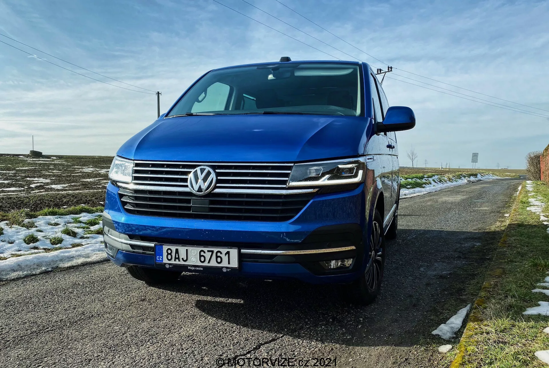 A Volkswagen T6.1 Multivan 2019-es modell alacsony, első háromnegyedes nézete, amelyen látható a vízszintes vonalakkal ellátott első rács, a rács közepén található jellegzetes VW embléma, a fényszórók, amelyek valószínűleg LED-es nappali menetfényekkel és bi-xenon vagy LED fényekkel rendelkeznek, valamint az első lökhárító ködlámpákkal és légbeömlőkkel. A kép a bal oldal egy részét is bemutatja, beleértve a vezető ajtaját és a sárvédőt, és látható a motorháztető és a szélvédő is. Az autó mély kék színű, és egy vidéki úton parkol, a földön hóval és felette tiszta kék égbolttal.