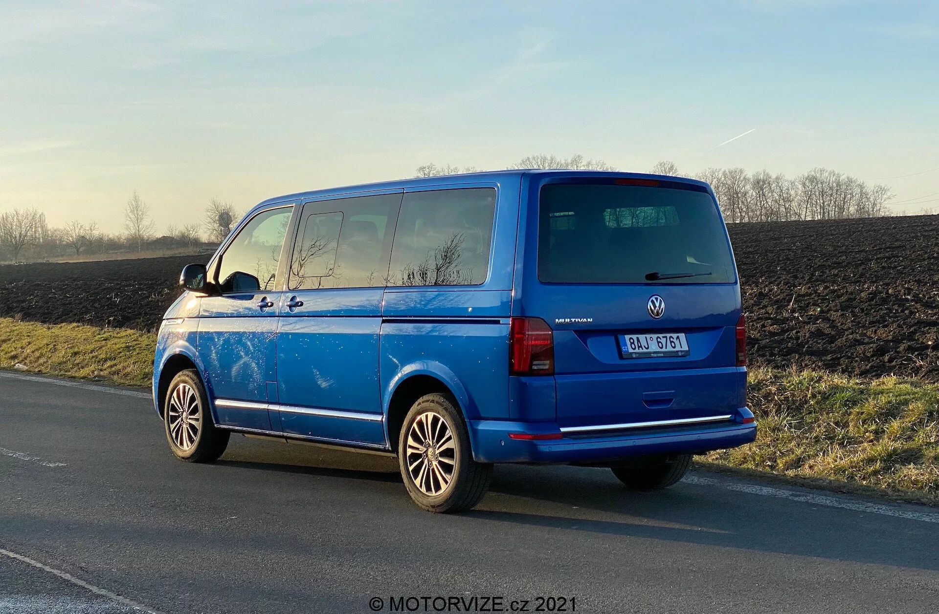 Élénk kék Volkswagen Transporter 2019 (T6-1 Multivan) parkol az út szélén, hátulról háromnegyedes nézetben, amely megmutatja a hátsó lámpákat, hátsó ablakot, hátsó ajtót, a gyártó jelvényét és a járműmodell részleteit; látható könnyűfém kerekek, külső oldalsó tükrök, hátsó ablakfűtés vonalai, sötétített másodlagos ablakok; tetőantennával ellátva; parkol természetes nappali fényben, a csillogó felület és a vidéki háttér kiemelésével.