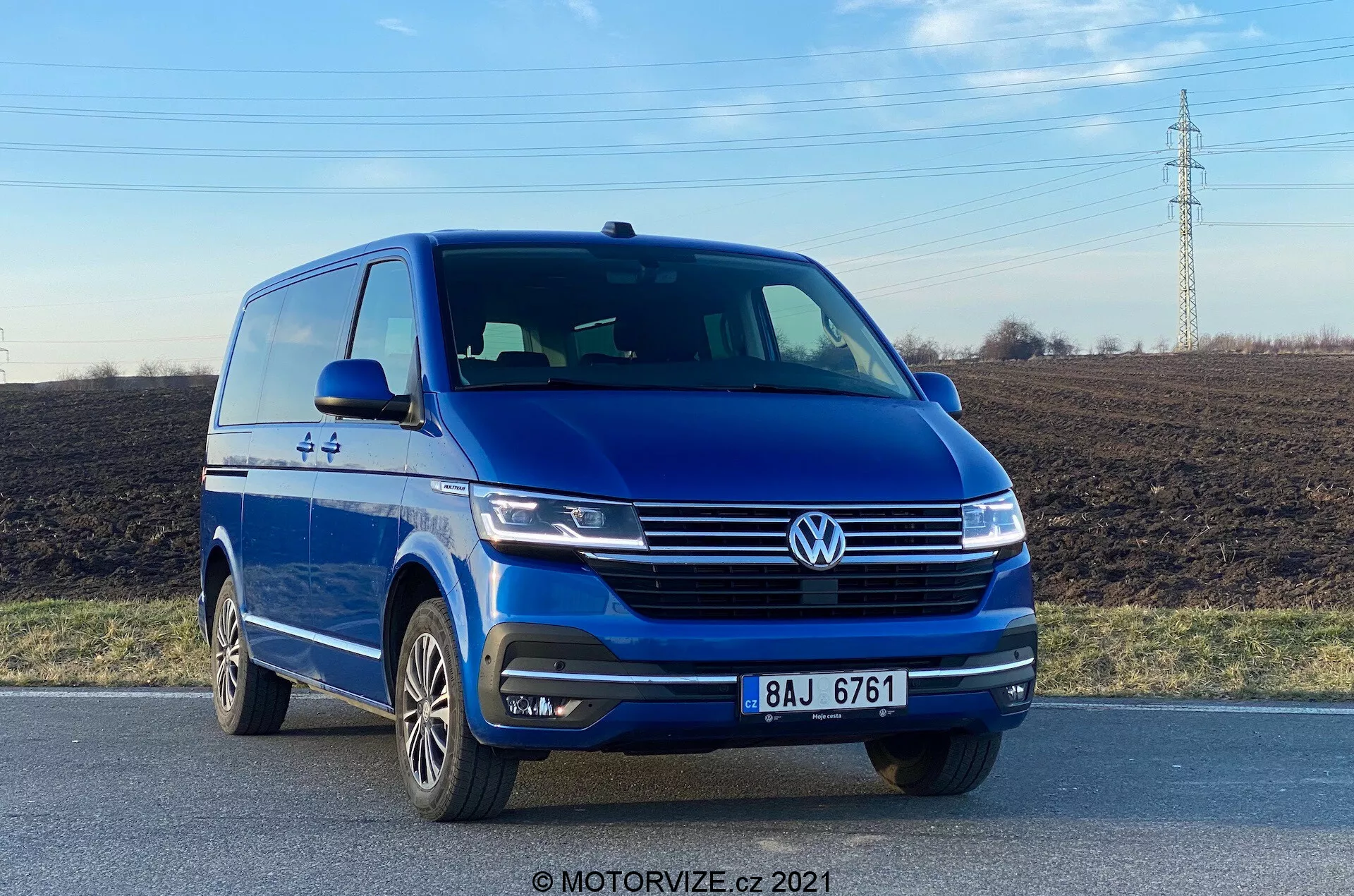 A kép egy kék Volkswagen VW-Nutzfahrzeuge Transporter 2019 (T6-1 Multivan) furgont ábrázol, alacsony frontális szögből fotózva, kiemelve a jármű bal első negyedét, világos kilátást nyújtva az orr részre és a bal oldali profilra. A jármű egy úton helyezkedik el, kiemelve az olyan elemeket, mint a hangsúlyos Volkswagen embléma, az elegáns fényszórók, a motorháztető formái, a lökhárítóba integrált ködlámpák és a többrészes alufelnik. A nappali fényben készült fotó hangsúlyozza a furgon külső megjelenését és a járművezetést segítő technológiákat, például a parkolásérzékelőket.