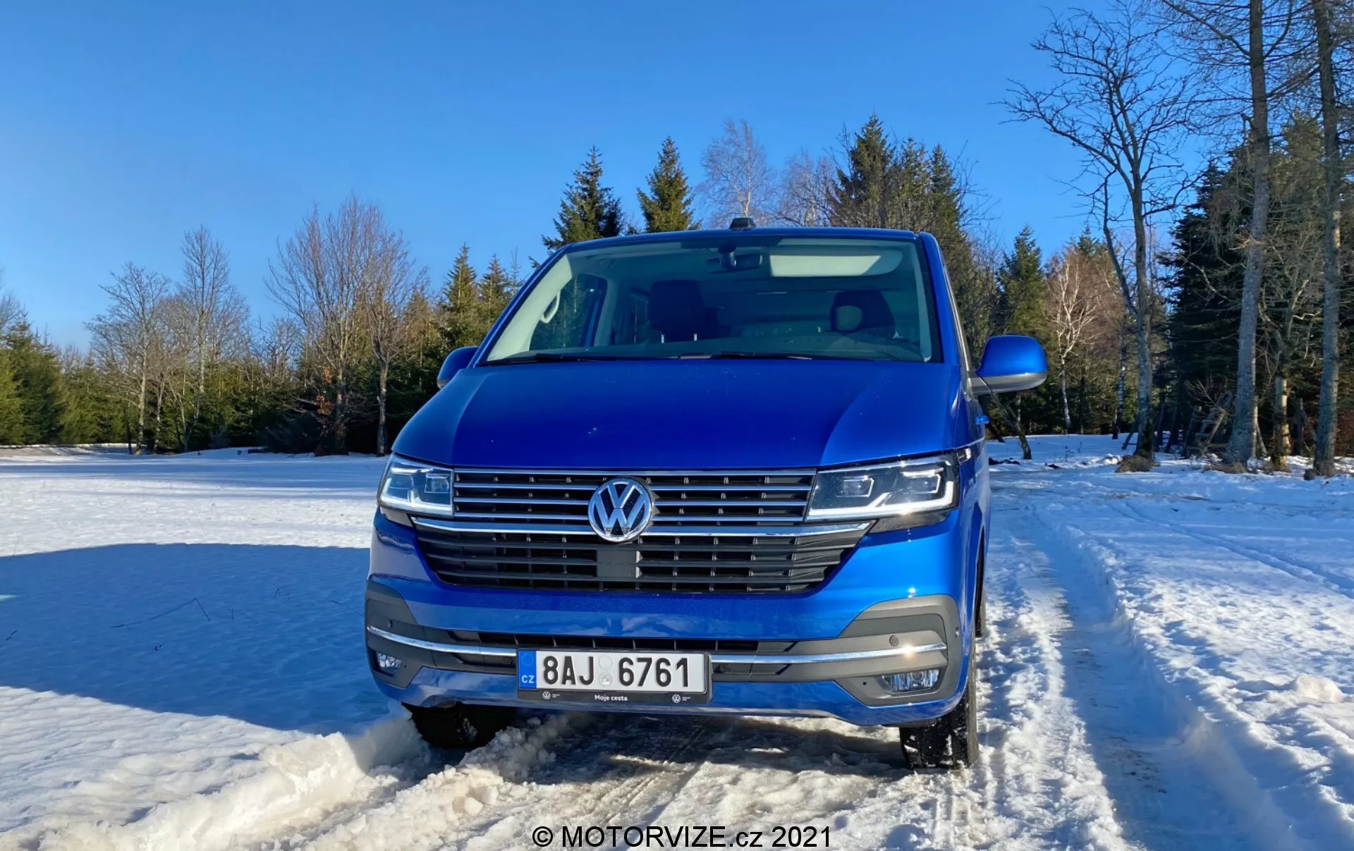 2019-es kék Volkswagen Transporter (T6-1 Multivan) elölnézeti fotója, amely a hűtőrácsot, fényszórókat és motorháztetőt mutatja. Hóval borított talajon elhelyezett jármű, tiszta égbolttal és fákkal a háttérben. Az autó jellemzői közé tartozik a VW embléma, vízszintes krómozott hűtőrács, LED fényszórók, elülső kamera, parkolásérzékelők, ezüst tetősínek és részben látható oldalsó tükör. Kortárs design aerodinamikus frontrésszel, nappali fotózás enyhe napfénnyel a jobb oldalról.