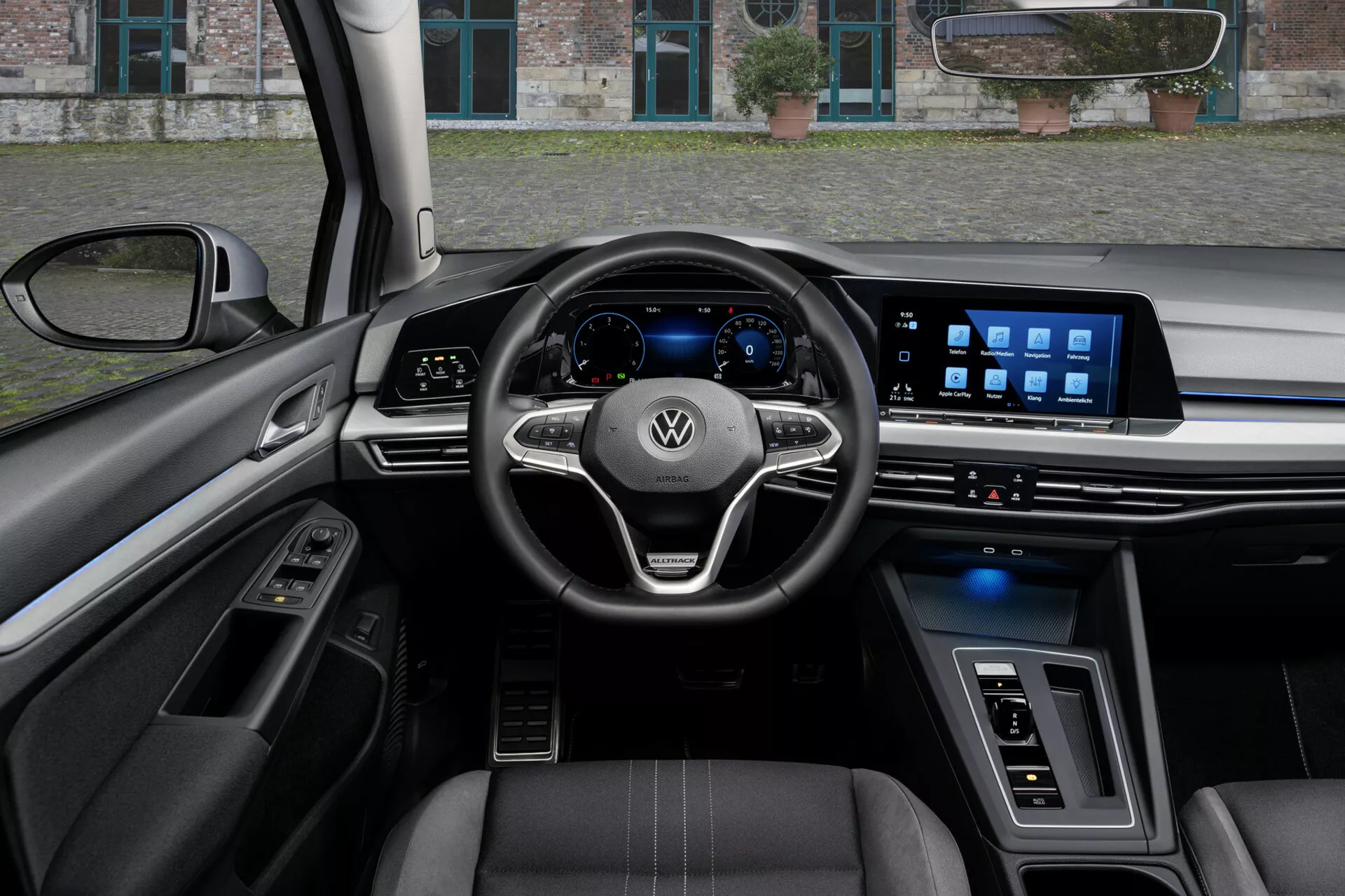 A 2017-es VW Golf Alltrack belsejének nézete a vezető perspektívájából, bemutatva egy háromküllős kormányt szerelvényvezérlőkkel és Volkswagen logóval, digitális műszerfalat és széles érintőképernyős infotainment rendszert a műszerfalon, kiemelve a fejlett technológiai jellemzőket, bőrrel borított üléseket kontrasztos varrással, és a luxus anyagok és ambient fények ötvözetét.