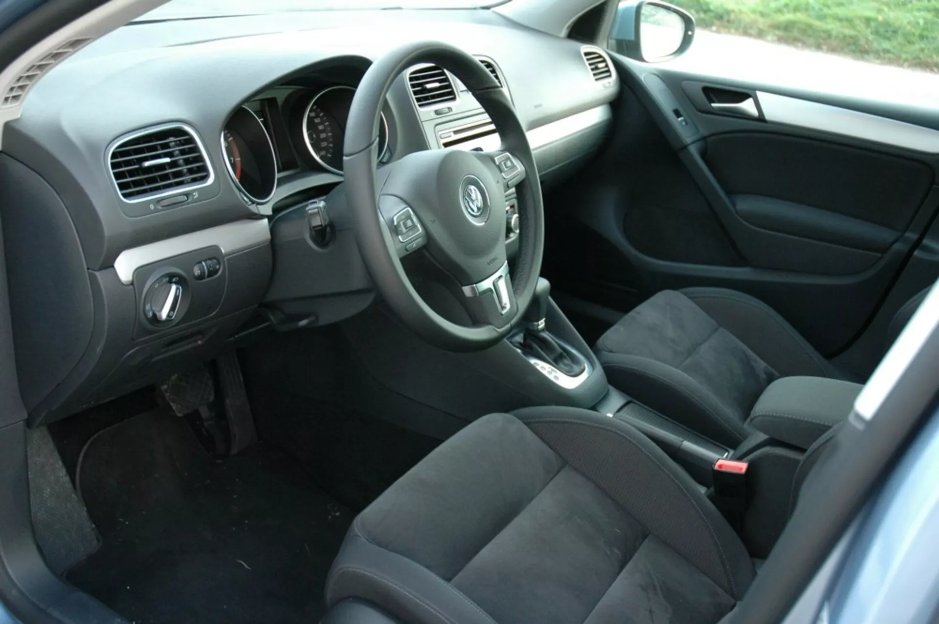 2008-as VW Golf belső terét mutató kép az utas oldalról a hátsó utas nézőpontjából az autó vezetői oldalával, a kormánnyal, műszerfalommal, középkonzollal, automatikus váltó karval, sötétszínű szövet ülésekkel, az vezető ajtajának vezérlőelemeivel, szőnyegezett padlóval, szőnyegekkel és természetes megvilágítással