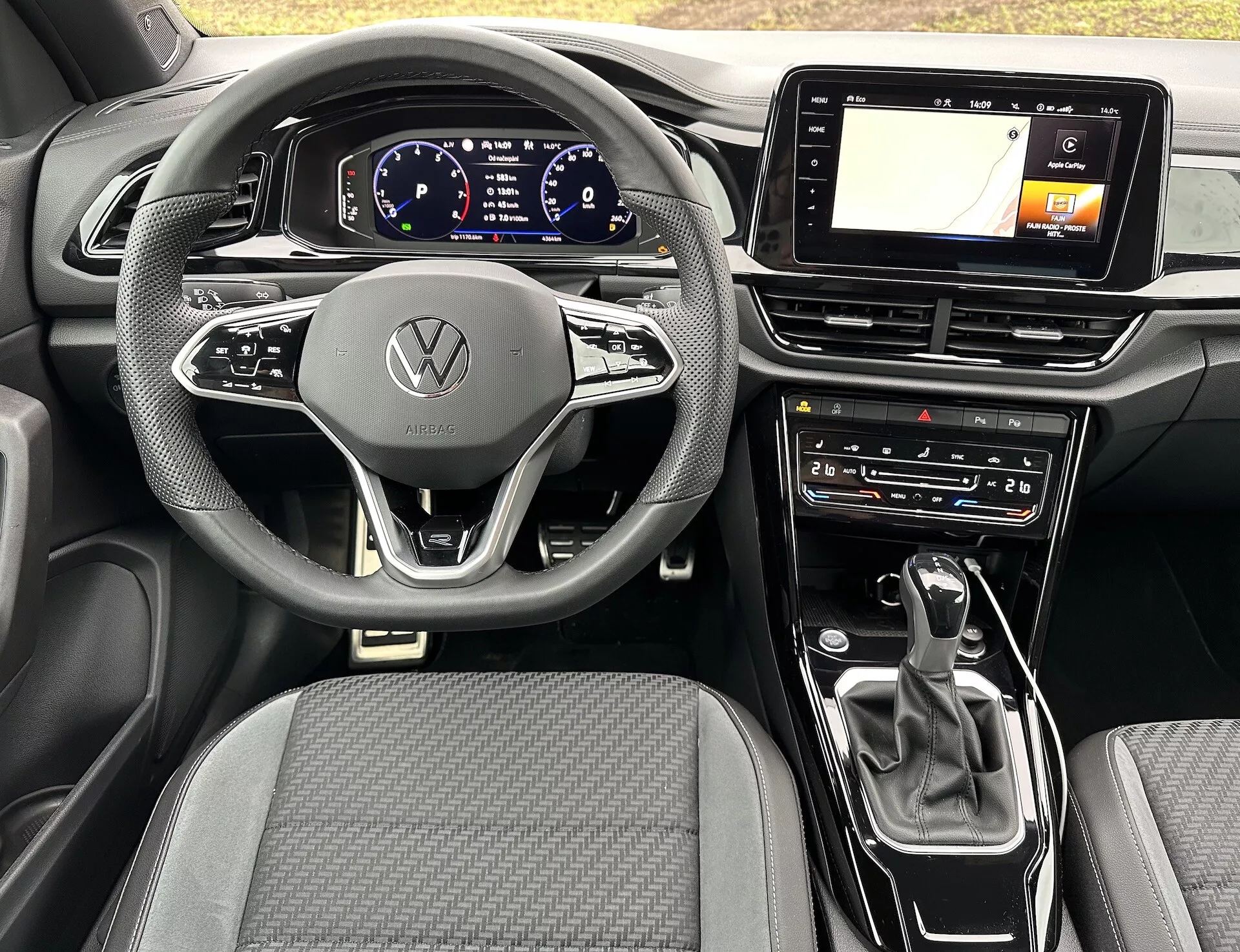 A 2022-es VW T-Roc Cabriolet belső nézete, amely egy modern háromküllős kormányt tartalmaz Volkswagen emblémával, digitális műszerfalat, GPS-képes infotainment érintőképernyőt, automatikus váltókarját és klímavezérlő beállításokat, kiemelve az autó fejlett technológiáját és letisztult dizájnját