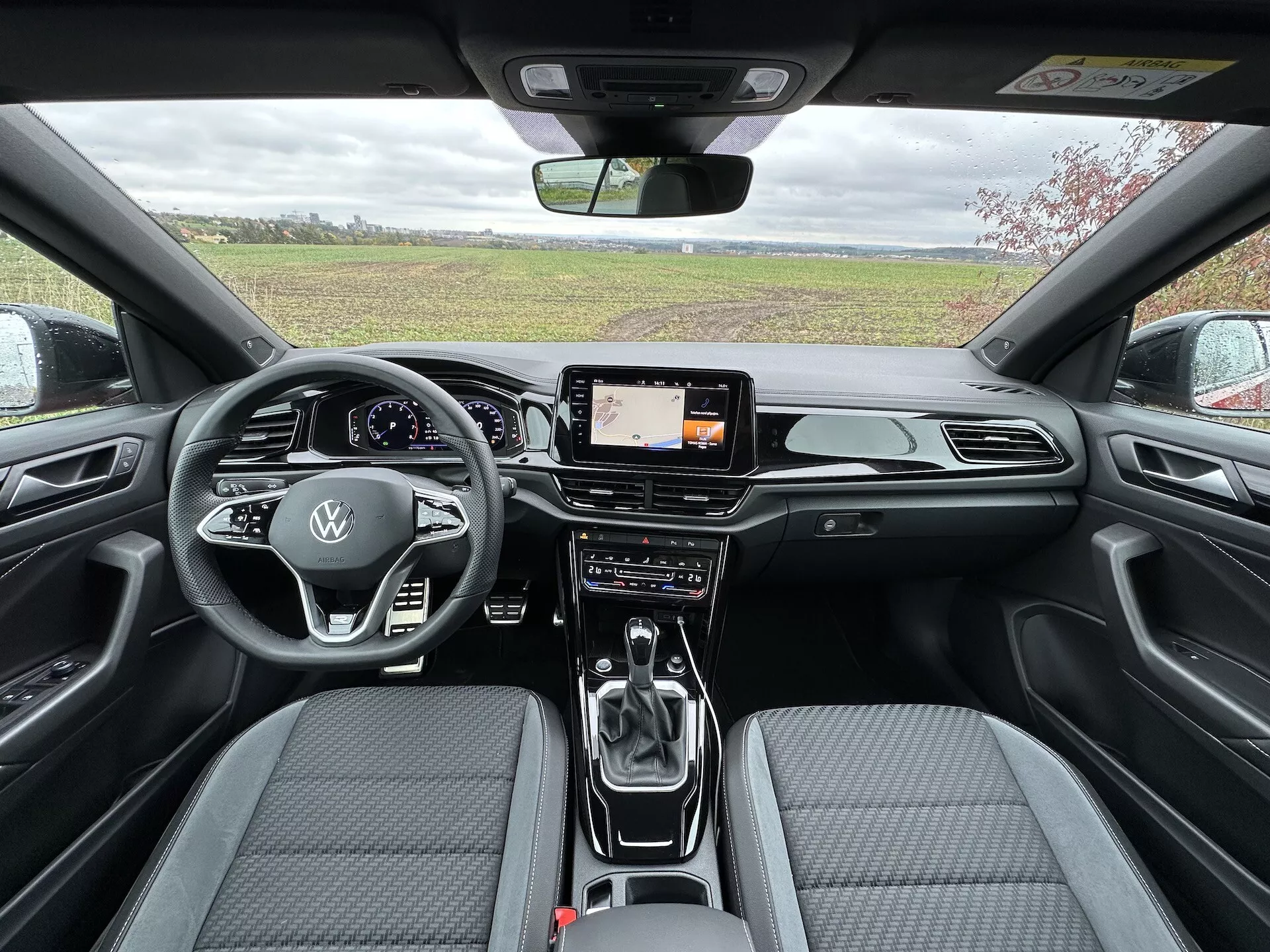 A 2022-es VW T-Roc Cabriolet belső nézete hátulról előre, amely a légterelő deszkát kromos díszítésekkel, digitális infotainment képernyőt, fejlett kettős zónás klímavezérlőt, bőrrel bevont multifunkciós kormánykereket Volkswagen logóval, automata váltókar, és kortárs kéttónusú szövet ülésekkel varrás mintával mutat be.