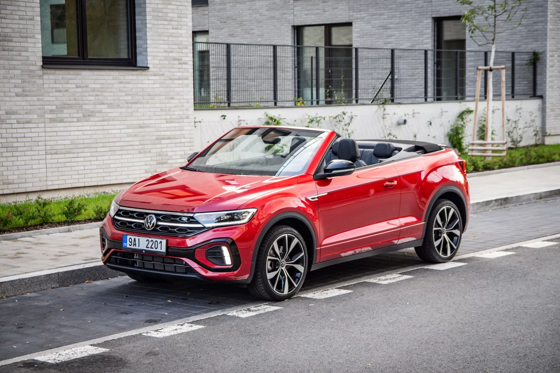 2022-es VW T-Roc Cabriolet parkol városi utcán, bemutatva az első hűtőrácsot, LED fényszórókat, lehajtott szövet kabrió tetőt és sportos többspókás kerekeket