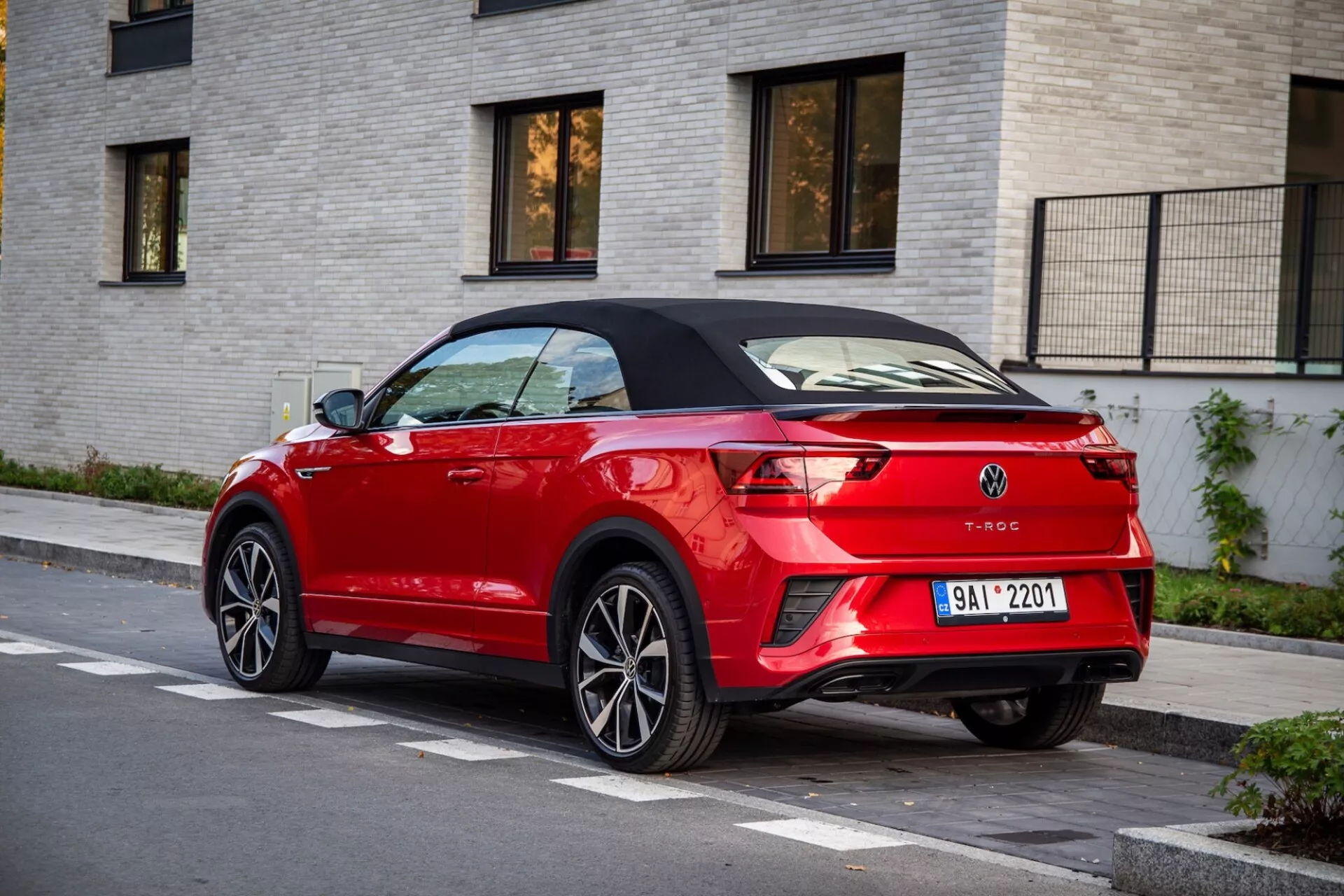 Vörös Volkswagen T-Roc Kabrió háromnegyed hátsó nézete parkolva egy utcán, kiemelve a fekete visszahúzható tetőt, a stílusos LED hátsó lámpákat, a hátsó spoiler ajkát, a kettős kipufogócsöveket és az integrált parkoló szenzorokat világos színű téglás épület hátterében.