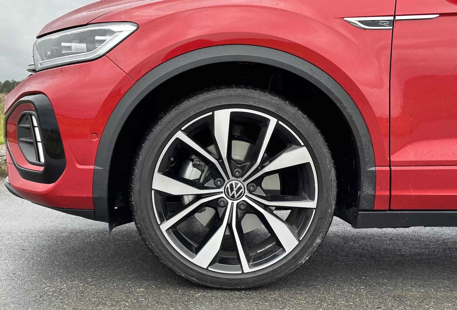 VW T-Roc 2022 közelképe, amely a többspókás ötvözött kerékre, kéttónusú befejezésre, alacsony profilú gumiabroncsra és a sportos vörös karosszéria és LED világítás részletezésére összpontosít a részleges első negyedpanelen