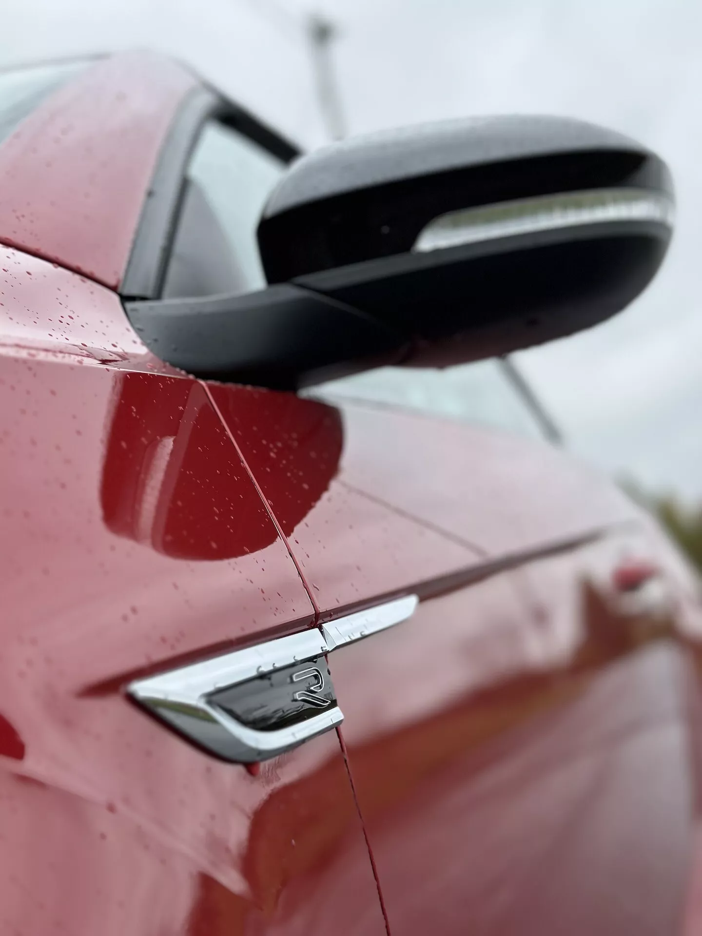 Közeli oldalsó nézet egy 2022-es piros VW T-Roc Cabriolet-re, kiemelve a jármű hátsó részét részletes megjelenítéssel a fekete oldalsó tükörről, kulcs nélküli belépést biztosító ajtókilincsről és a jármű tervezési elemeiről, beleértve a jelvényt, enyhén felfelé irányuló szög alatt vízcseppekkel a felületen, jelezve friss esőt vagy mosást, elmosódott háttérrel