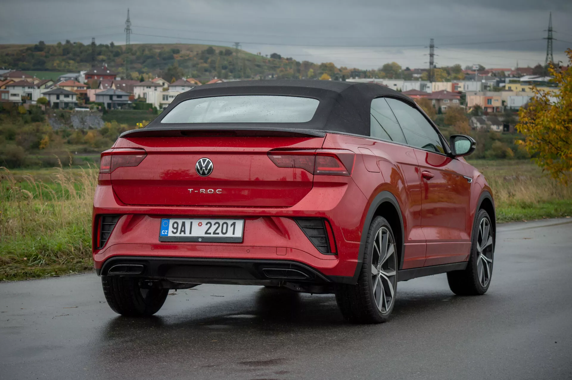 Vörös Volkswagen T-Roc Cabriolet 2022 háromnegyedes hátsó nézetben nedves úton, bemutatva a hátsó dizájnt, oldalnézetet, keréktárcsák dizájnját, LED hátsó lámpákat, beépített érzékelőkkel rendelkező hátsó lökhárítót, kettős kipufogócsöveket, többsugaras ötvözött kerekeket és a kabrió tetőt
