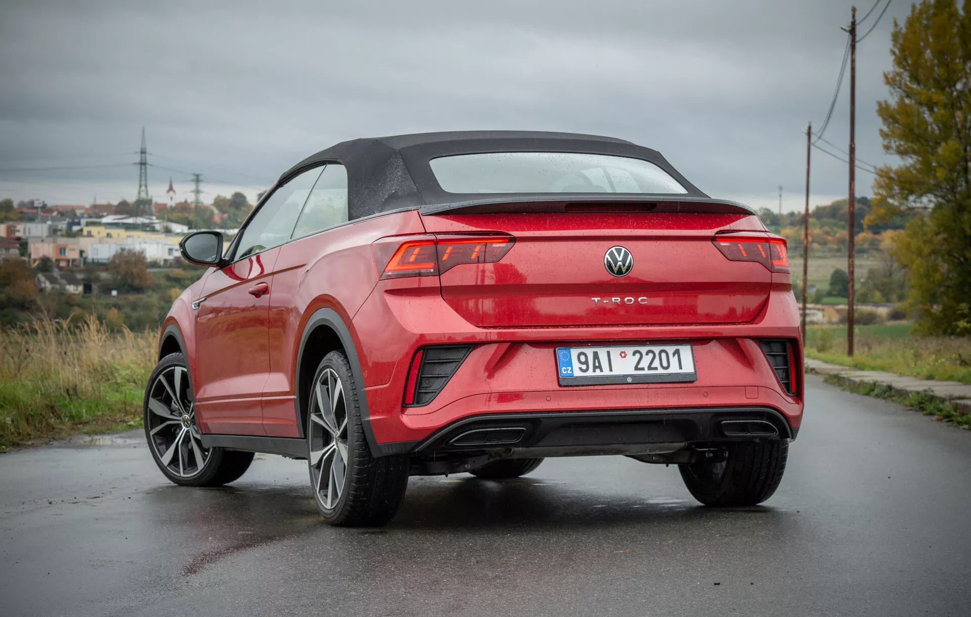 Háromnegyedes hátsó nézetű 2022-es VW T-Roc Cabriolet parkol egy nedves úton, bemutatva a modern kabrió tervezését, éles karosszéria-vonalakat, sportos ötvözött kerekeket, elegáns hátsó lámpákat és kettős kipufogót, félvidéki háttérrel, elektromos oszlopokkal és látványos zöldövezettel.