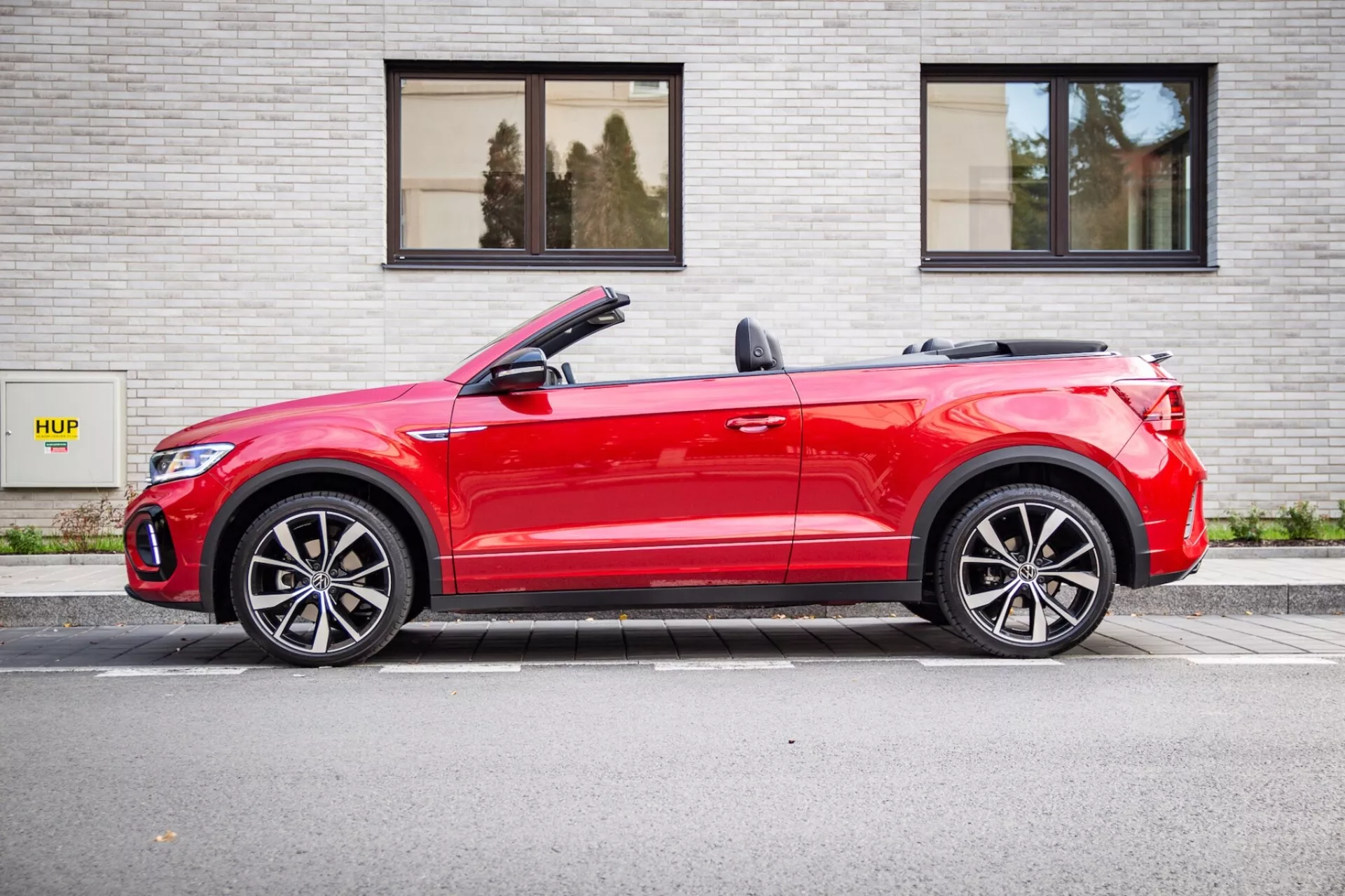 2022-es vörös Volkswagen T-Roc Cabriolet, párhuzamosan parkolva egy szürke téglafal mellett HUP táblával, oldalprofil látható enyhén az elejéből, lehajtott kabriótető, LED fényszórók, és sportos ötvözött kerekek
