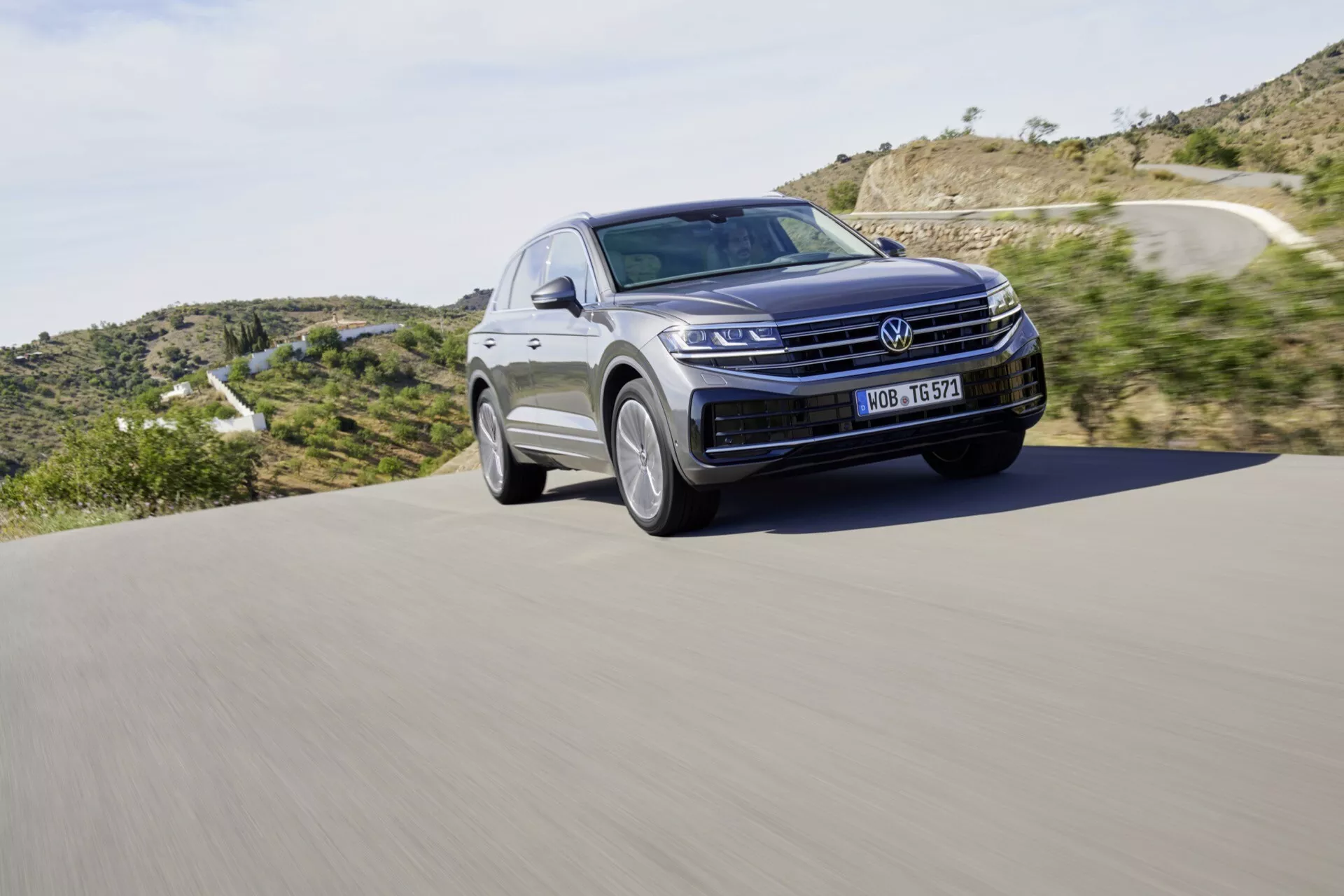 2023-as Volkswagen Touareg III Facelift SUV mozgásban egy aszfaltozott úton, kiemelkedő első ráccsal, LED fényszórókkal, és a motorháztető sima vonalával egy természetes táj hátterével, hangsúlyozva a modern dizájnt és autóipari technológiát.