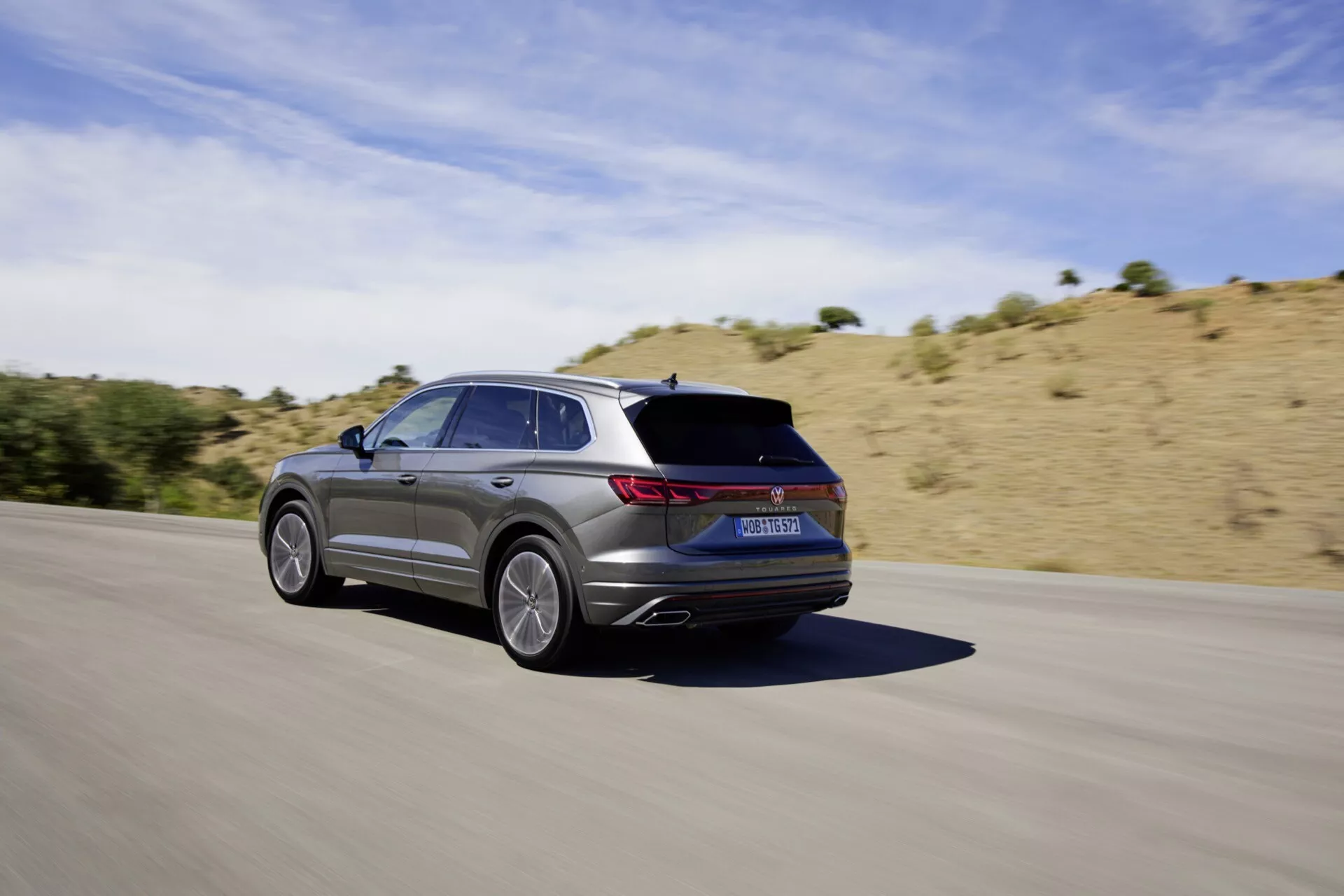 2023-as VW Touareg III facelift mozgásban az aszfaltúton, hátulról háromnegyedes szögből felvéve, bemutatva az elegáns kialakítását, ötágú ötvözött kerekeit, fémes bevonatát, LED hátsó lámpáit, integrált kipufogóit és sportos esztétikáját száraz, dombos táj ellenére tiszta napsütéses égbolt alatt